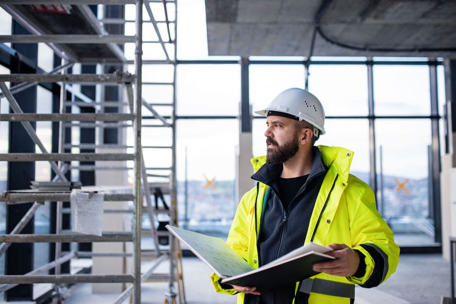 Construction worker in hi-vis yellow jacket and white hard hat holding a clipboard/notebook on a construction site